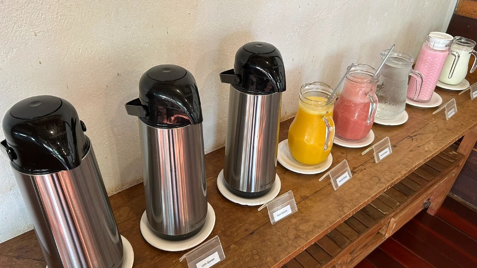 Uma fileira de distribuidores de bebidas em aço inoxidável, cada um com tampas pretas e uma etiqueta com o nome.