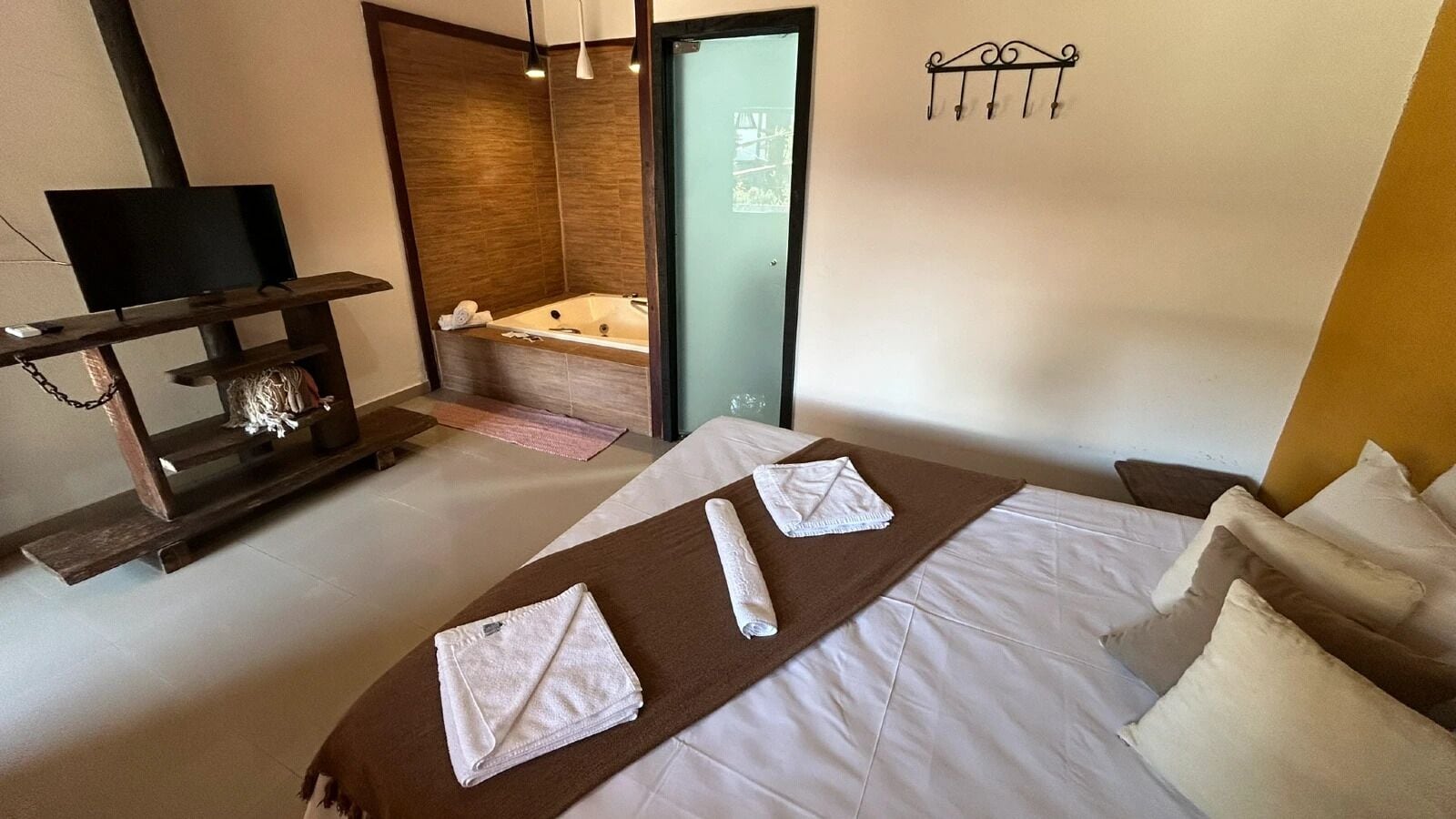Habitación de hotel con cama, televisor, una mesita de noche con lámpara, bañera y toallero de pared.