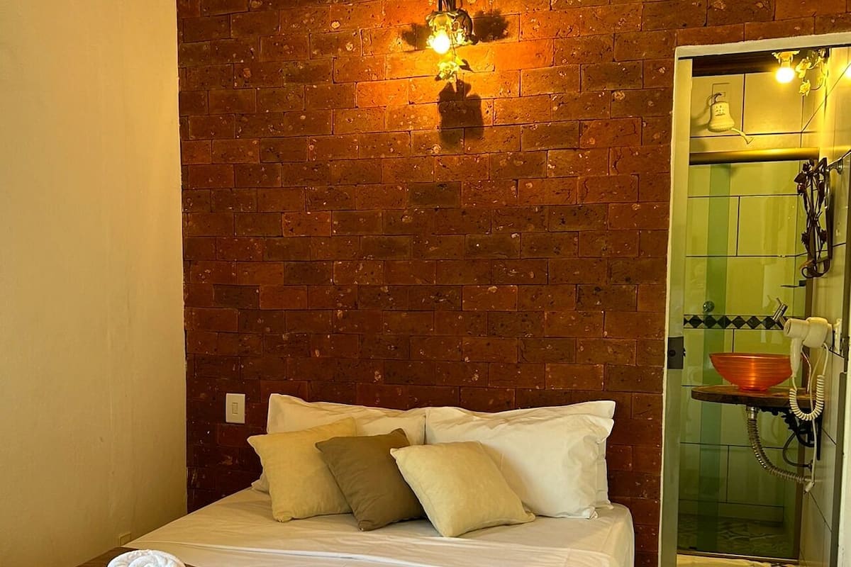 A room with a brick wall, a bed with pillows, a hanging light fixture, and a bathroom with a sink and mirror.
