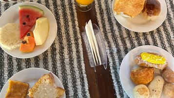 Mesa de café da manhã com pratos de frutas, torradas e pães doces sobre toalha de tecido trançado.
