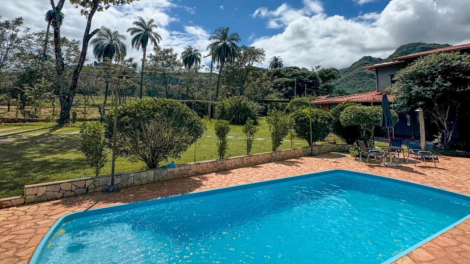 Una piscina con borde de piedra, rodeada por un patio de piedra, y vistas a un jardín exuberante con palmeras y una montaña al fondo.