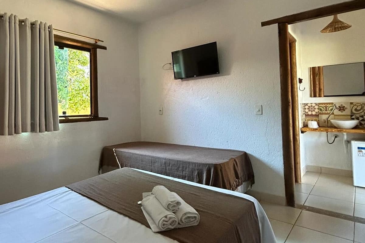 A room with two beds, a television on the wall, a window with curtains, and a bathroom with a sink and mirror.