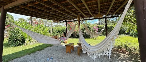 Una hamaca bajo una pérgola de madera, rodeada por un área de grava y abundante vegetación.