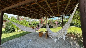 Una hamaca bajo una pérgola de madera, rodeada por un área de grava y abundante vegetación.
