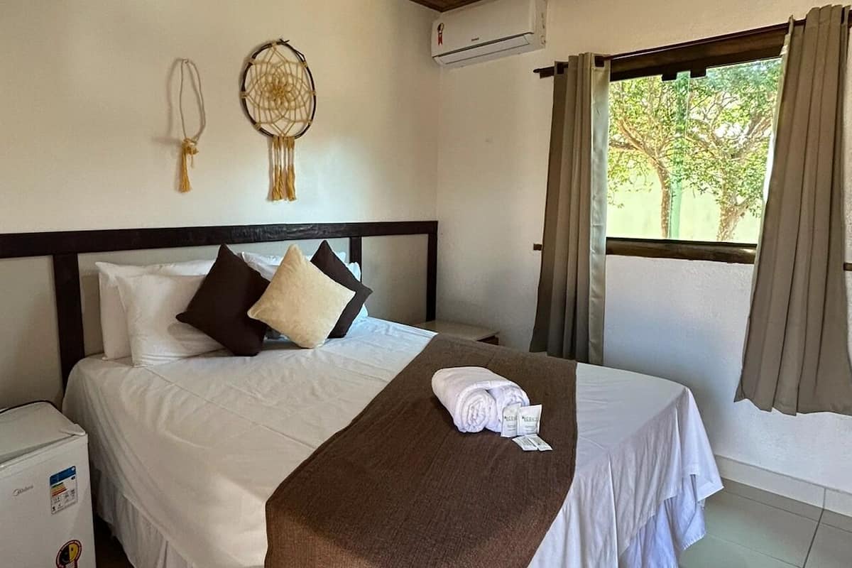 A hotel room with a bed, a bedside table with a towel and toiletries, a wall-mounted air conditioner, and a window with curtains.