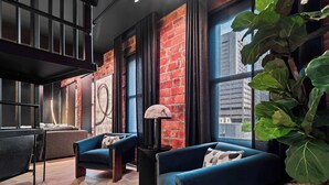 A modern living room with a brick wall, a large window, a sofa, a chair, a lamp, and a potted plant. - RoofTopPatio–HotTub–FirePit-WetBar-Downtown (Cleveland)