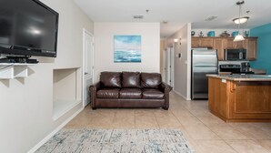 A modern living room with a flat-screen TV, a brown leather sofa, a kitchen with wooden cabinets, a stainless steel refrigerator, and a blue accent wall. - Star Beachside - Marlin Cottages 1 - Beautiful Condo, Family Friendly, Swimming & Kiddie Pool, Spa, Short Walk to the Beach (South Padre Island)