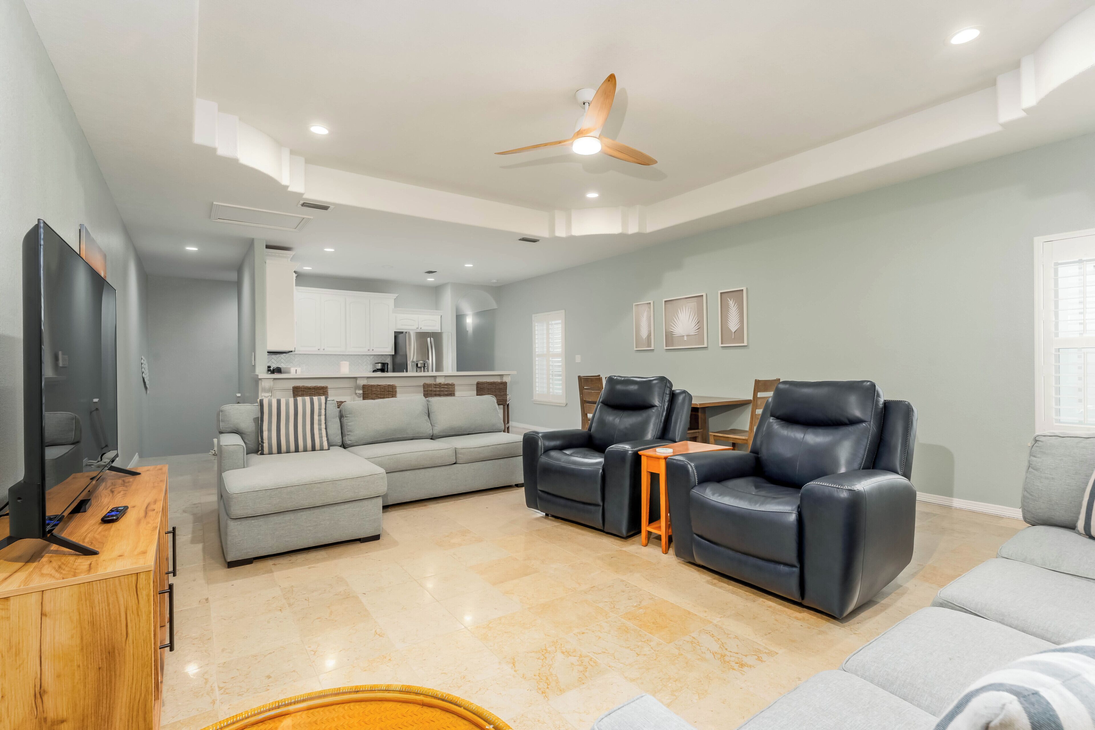 A spacious living room with a sofa, two armchairs, a coffee table, a TV, and a ceiling fan.