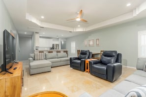 A spacious living room with a sofa, two armchairs, a coffee table, a TV, and a ceiling fan.