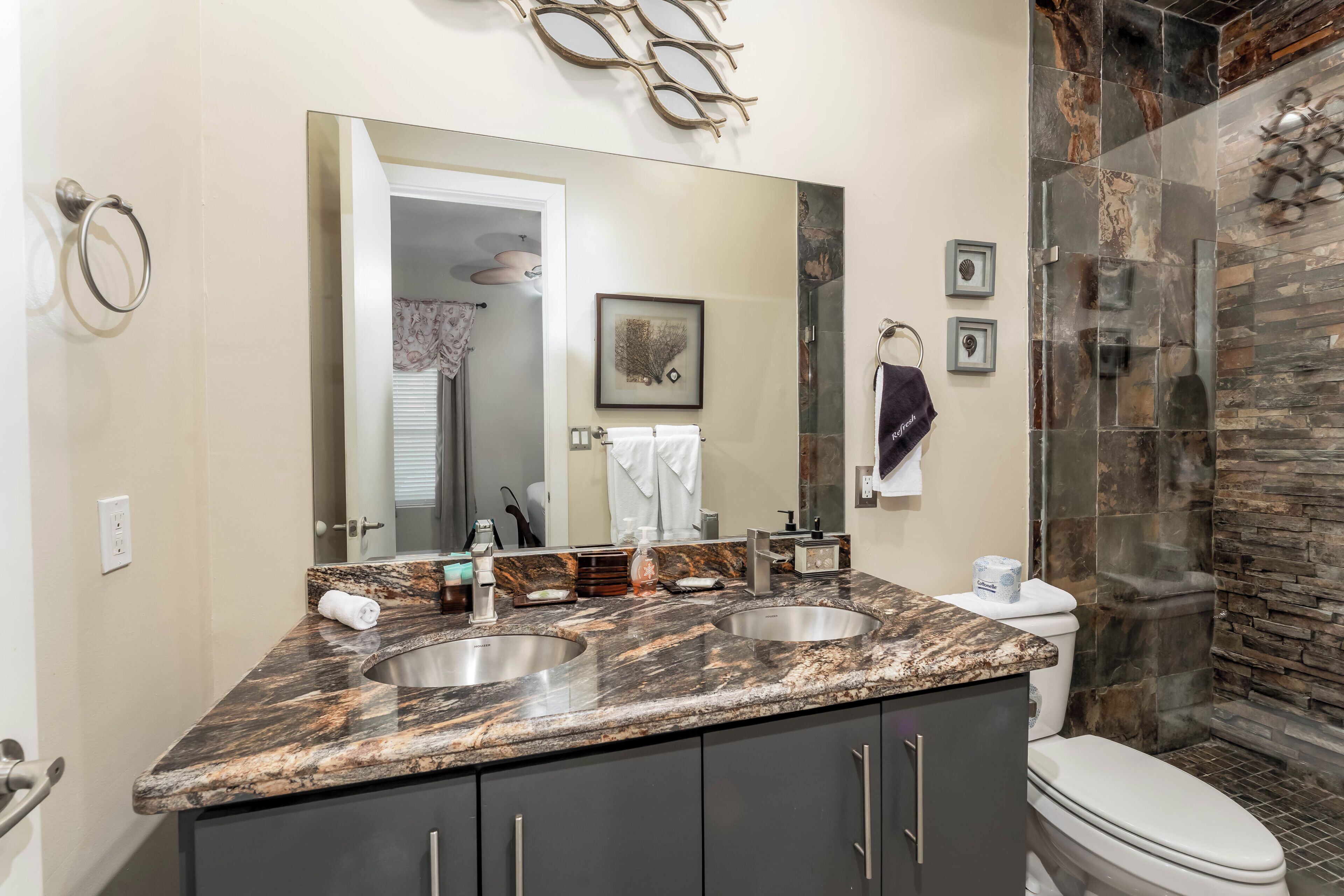 Une salle de bain dotée d’un comptoir en marbre, de deux lavabos, de toilettes, d’un grand miroir et d’une douche aux carreaux en pierre.