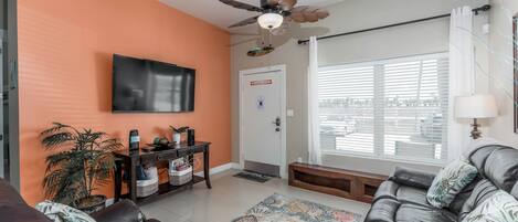 Una sala de estar con sofá, televisor, ventilador de techo, ventana con persianas y puerta.