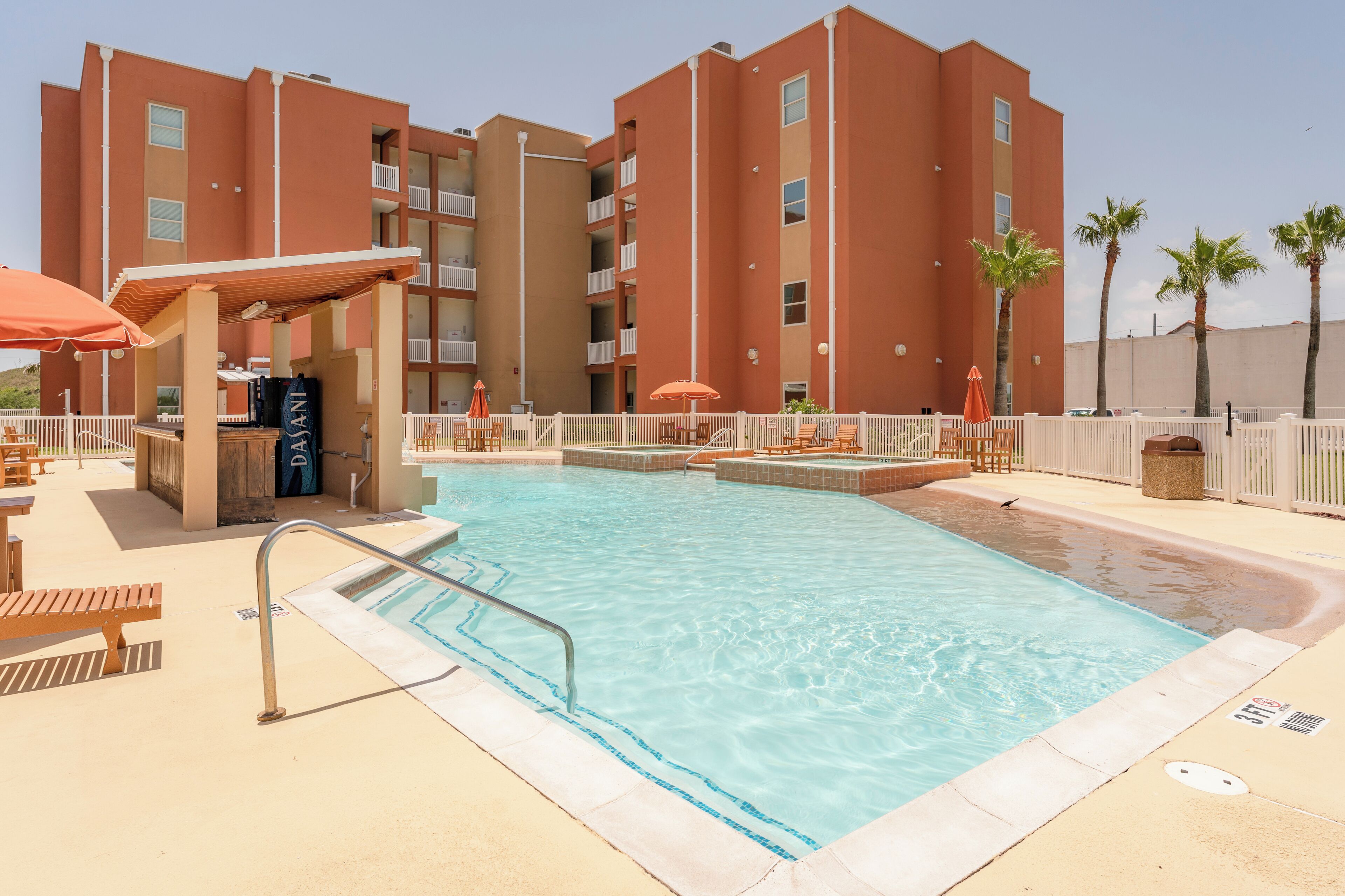 A swimming pool with a sunshade, lounge chairs, and a building with balconies.