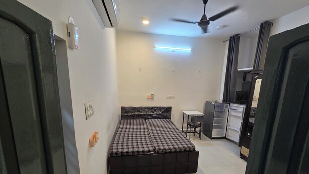 A compact bedroom with a bed, a small table, a chair, a ceiling fan, and a refrigerator. - Peaceful 1BHK 🌿 WiFi near by GT Mall, Airport” ✅ Jawahar circle malviya nagar (Jaipur)