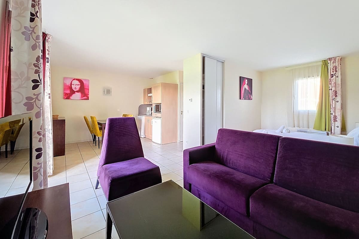 A hotel room with a purple sofa, a dining table with yellow chairs, a kitchen area, and a bed with white bedding.