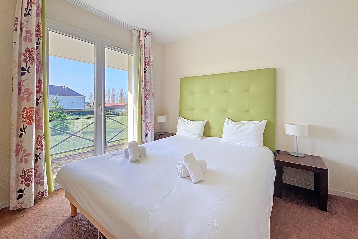 A hotel room with a large bed, two pillows, a green headboard, a nightstand with a lamp, and a view of a building and greenery outside.