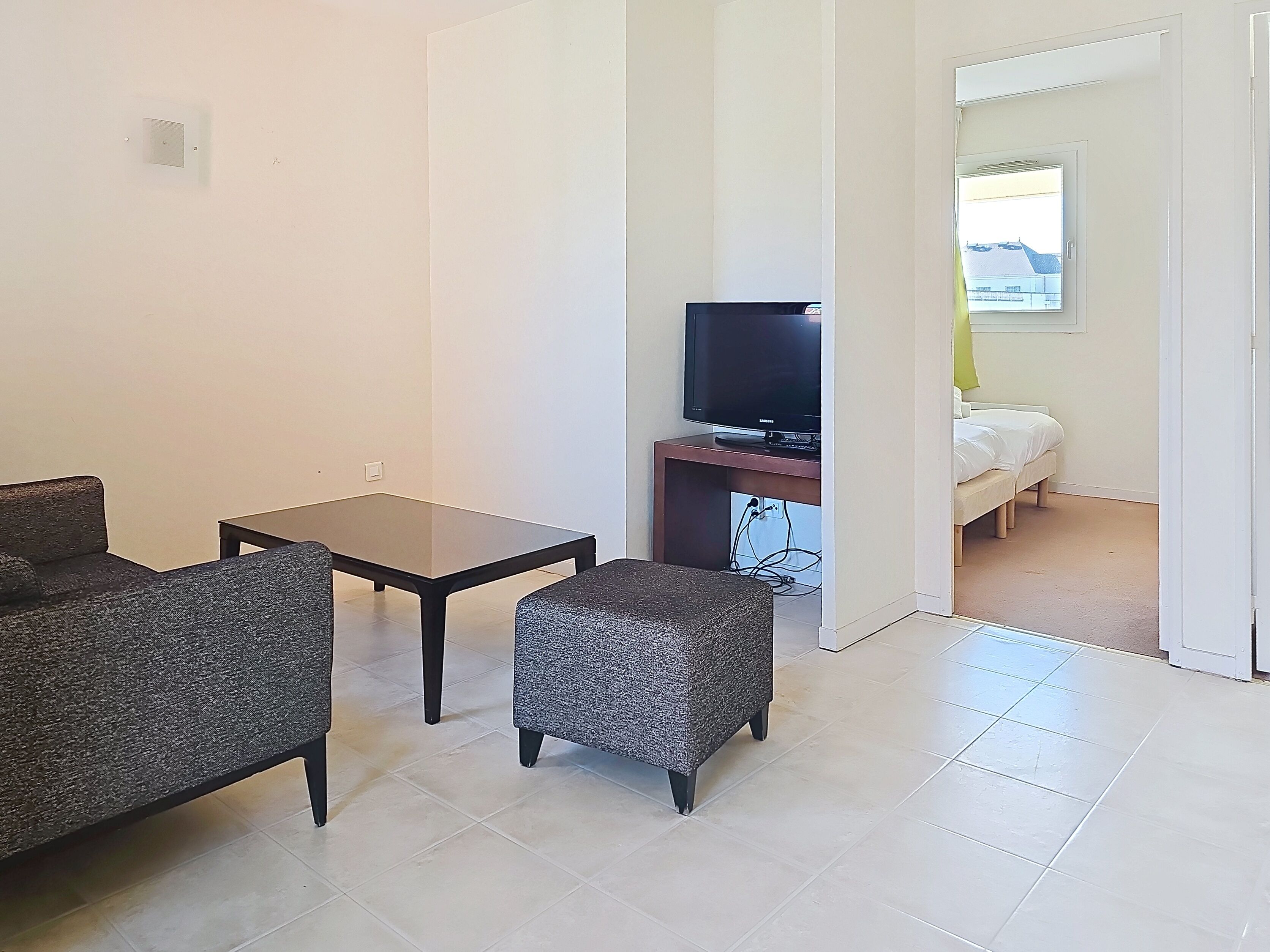 Ein Zimmer mit einer Couch, einem kleinen Tisch, einem Fernseher auf einem Schrank und einem Bett im Hintergrund.