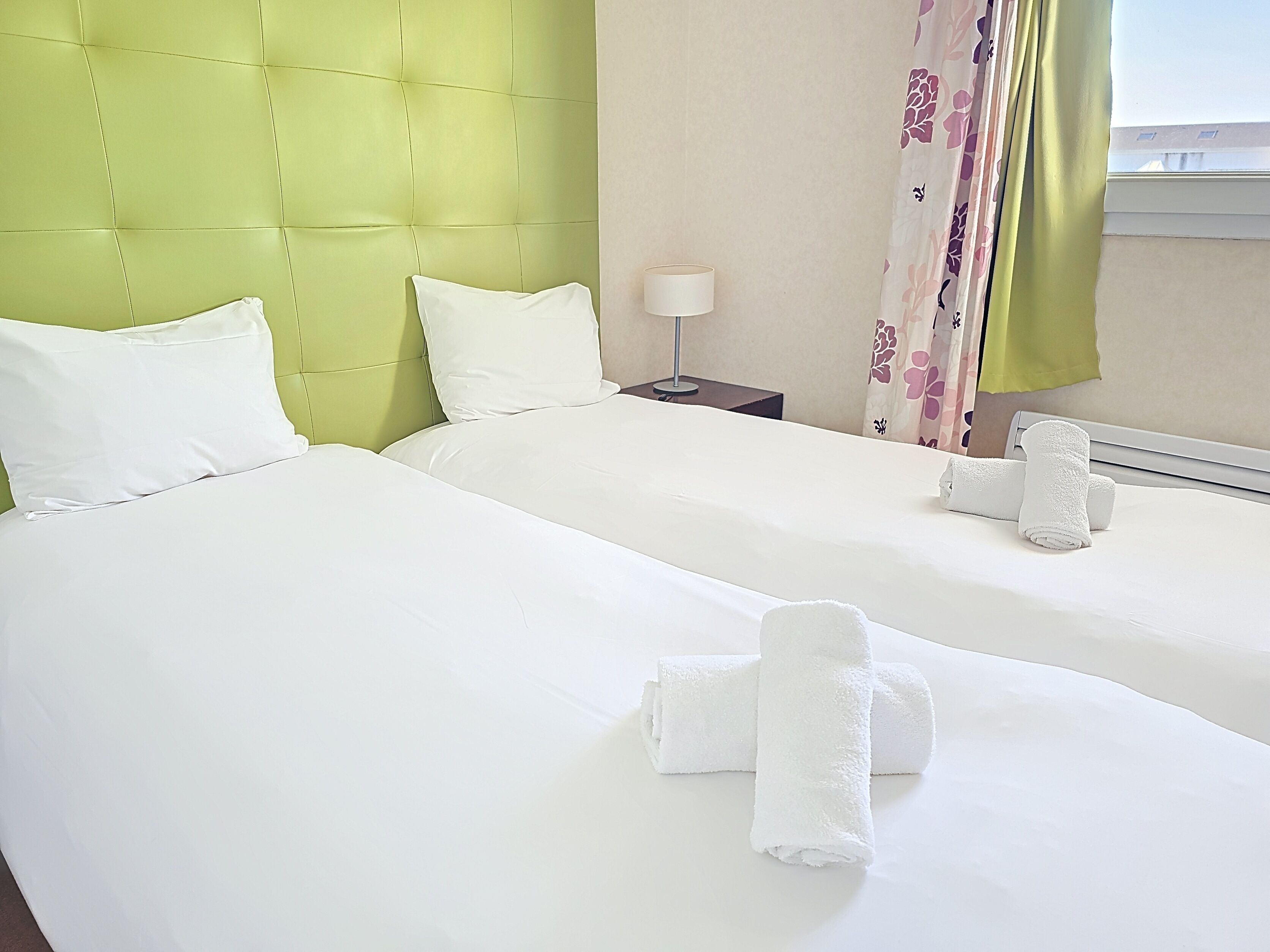 A hotel room with two beds, a green headboard, white bedding, a lamp, and a window with floral curtains.