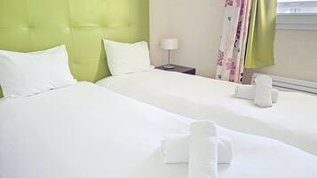 A hotel room with two beds, a green headboard, white bedding, a lamp, and a window with floral curtains.