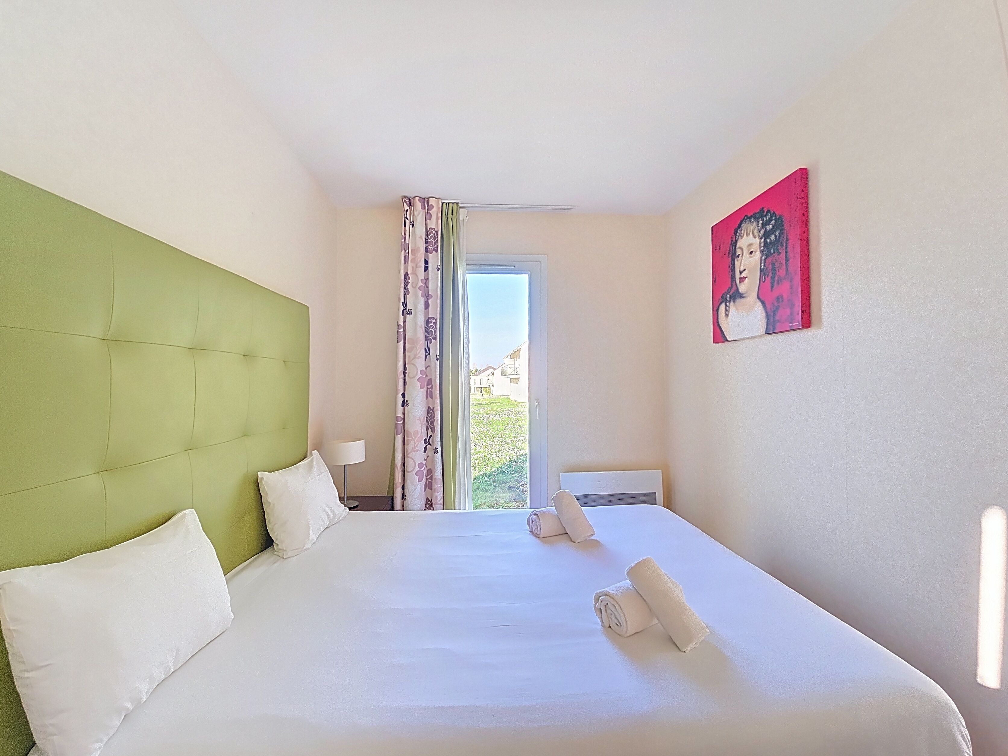 Une chambre d’hôtel avec une tête de lit verte, un lit avec du linge de maison blanc, un tableau au mur et une fenêtre avec des rideaux.