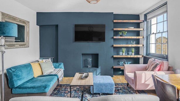 Un salón con sofá, mesa de centro y televisor montado en la pared. Se observan estanterías con plantas y una ventana con persianas.