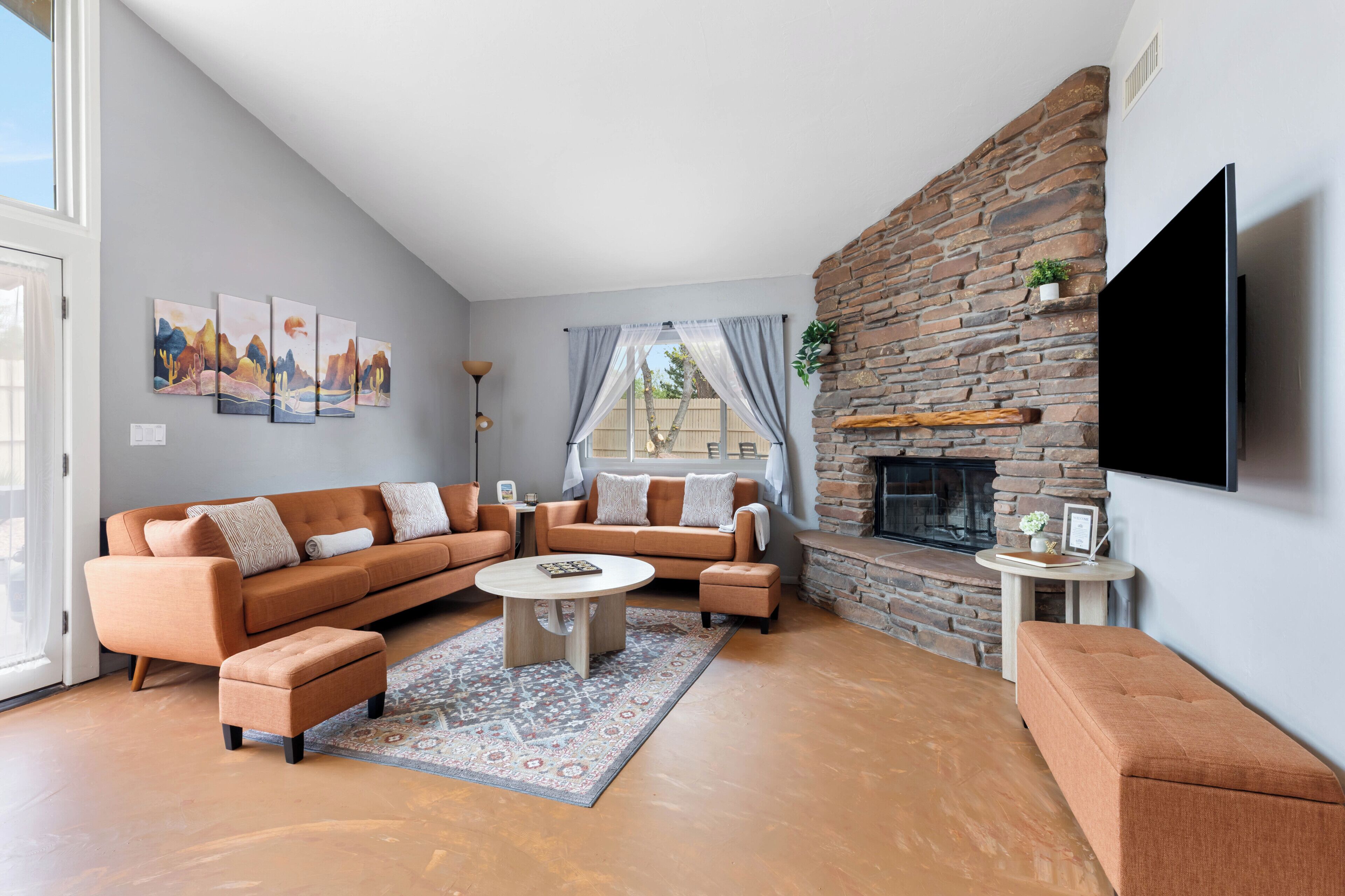 A living room with a sectional sofa, ottoman, coffee table, fireplace, flat-screen TV, artwork, and a window with curtains.