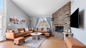 Ein Wohnzimmer mit Ecksofa, Hocker, Couchtisch, Kamin, Flachbildfernseher, Kunstwerken und einem Fenster mit Vorhängen.