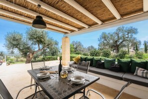 Un espace de restauration en plein air avec une table dressée pour deux personnes, une bouteille de vin et une vue sur des oliviers et la verdure.