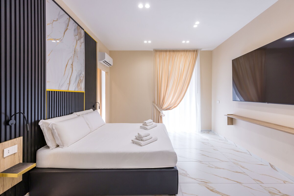 A modern hotel room with a large bed, a flat-screen TV, a built-in air conditioning unit, and a large window with curtains.