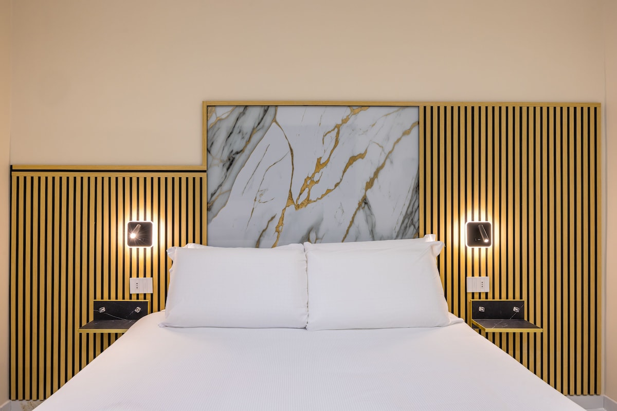A hotel room with a bed, a marble headboard, vertical wooden paneling, and bedside tables with lamps.
