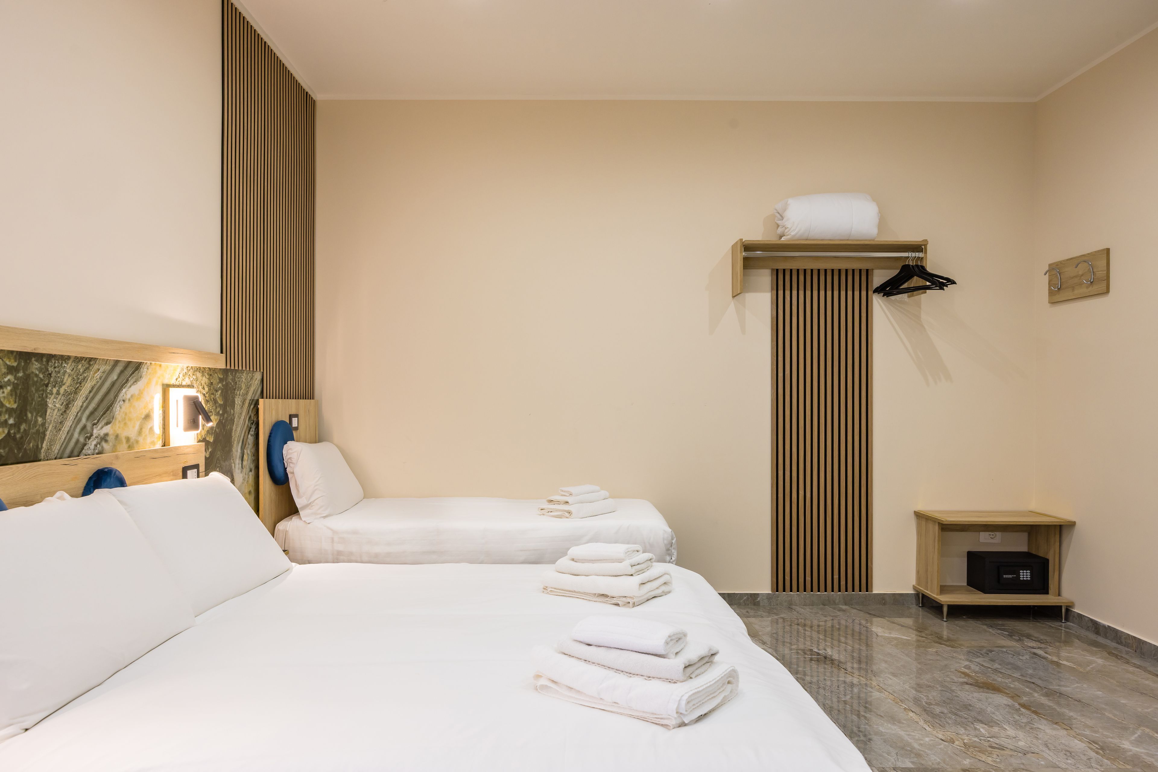 Habitación de hotel con dos camas, un estante de madera con toallas, una mesita de noche y un perchero para toallas en la pared.