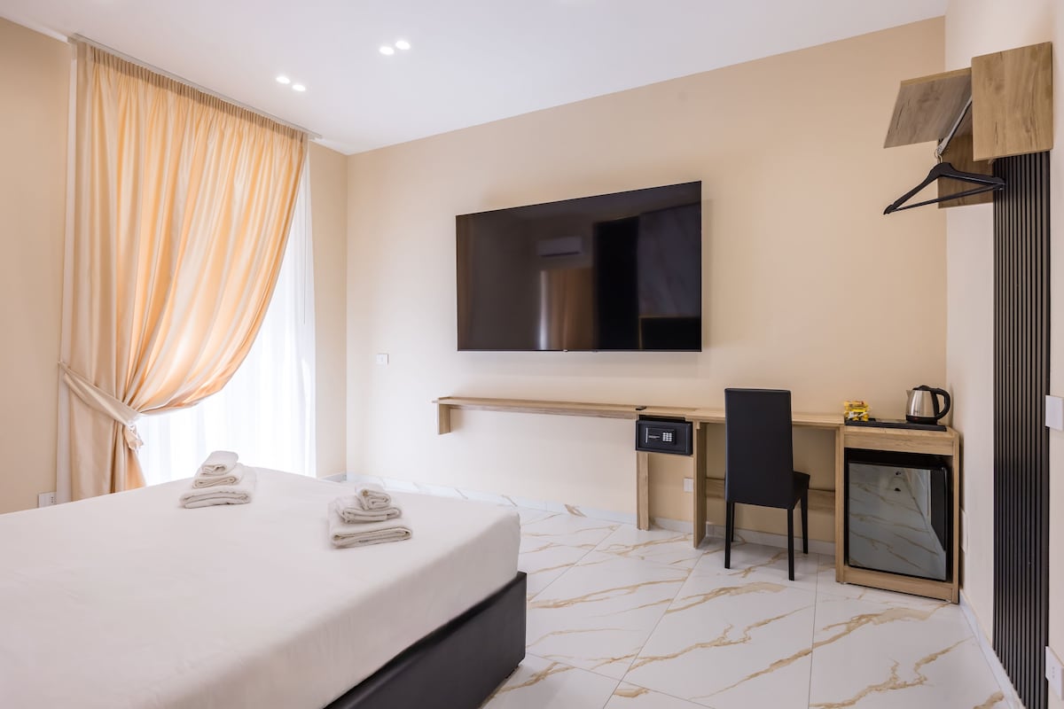 A hotel room with a bed, a desk, a chair, a television, and a small refrigerator.
