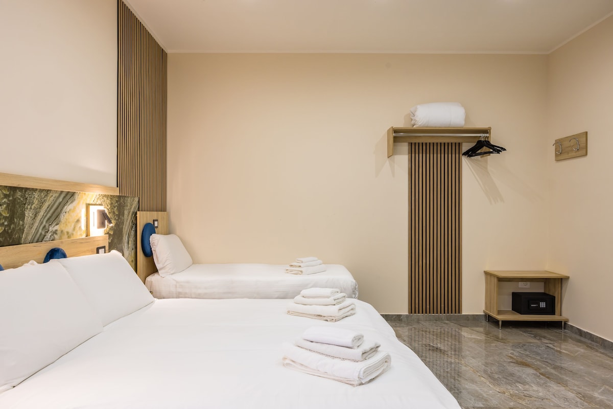 A hotel room with two beds, a wooden shelf with towels, a small bedside table, and a wall-mounted towel rack.