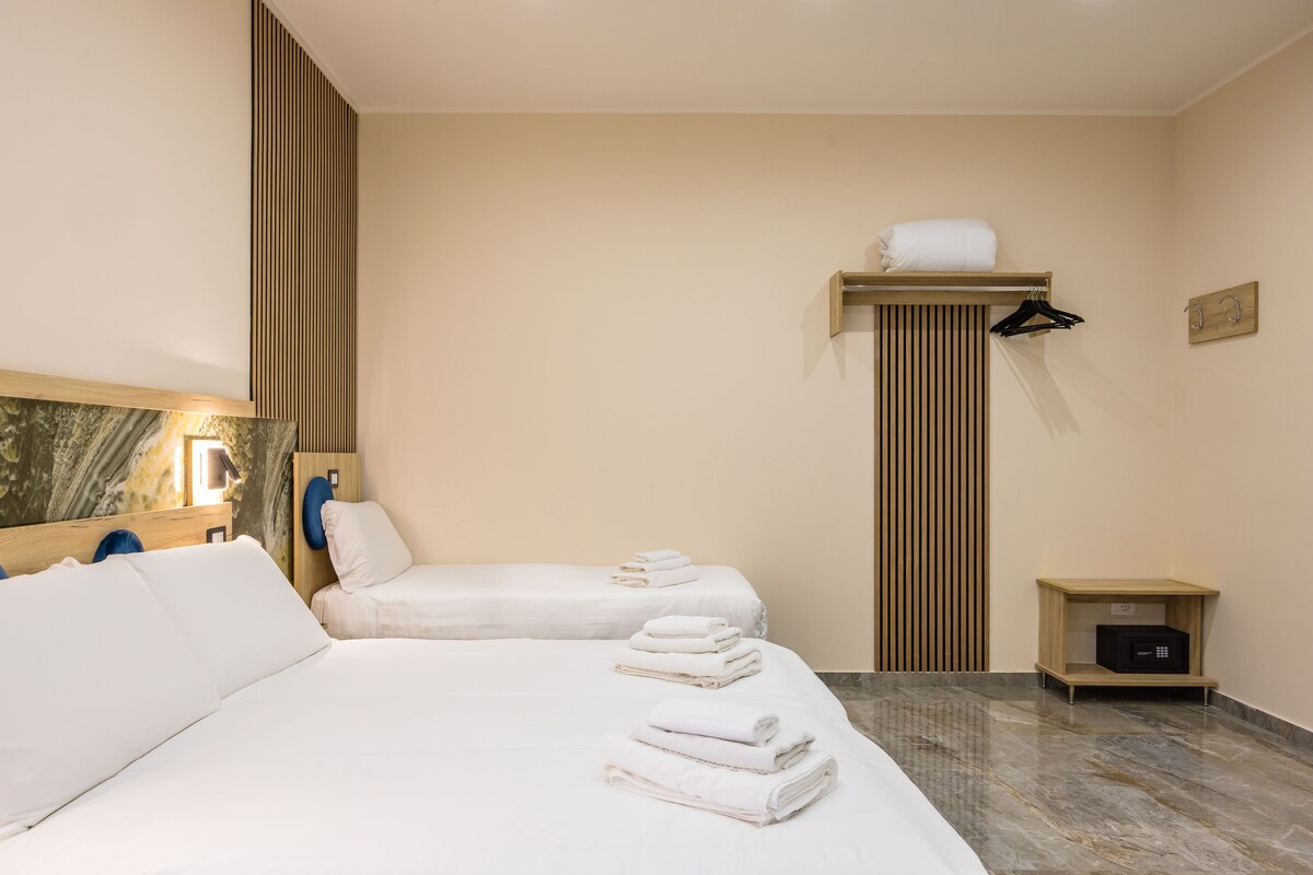 A hotel room with two beds, a wooden shelf with towels, a small bedside table, and a wall-mounted towel rack.