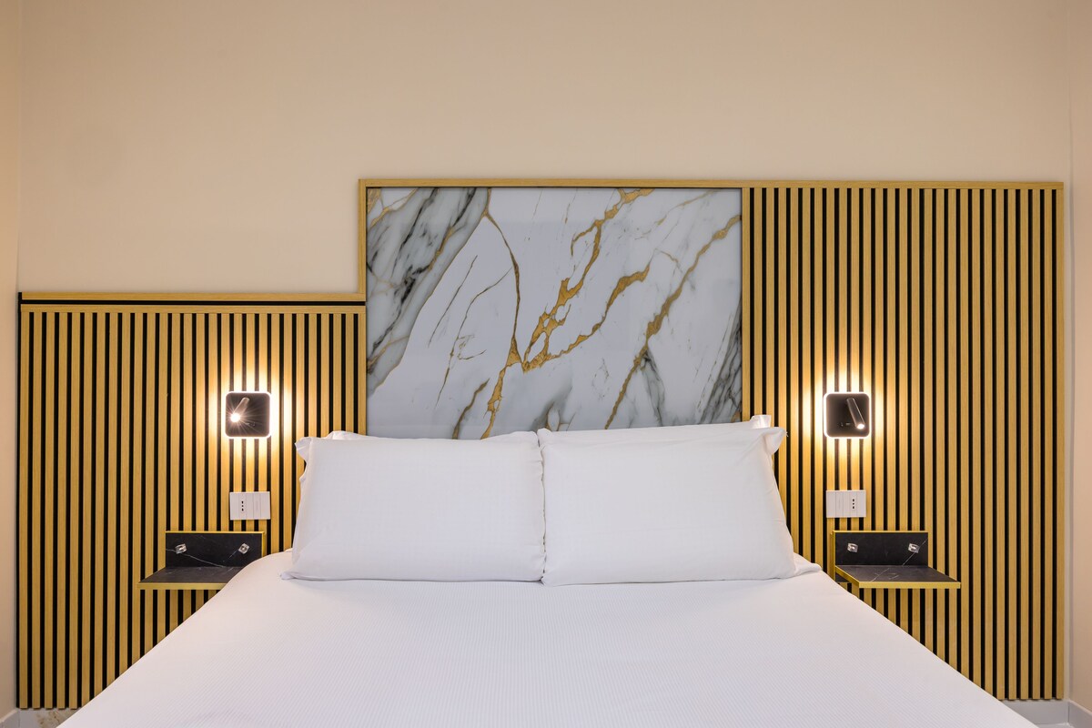 A hotel room with a bed, a marble headboard, vertical wooden paneling, and bedside tables with lamps.