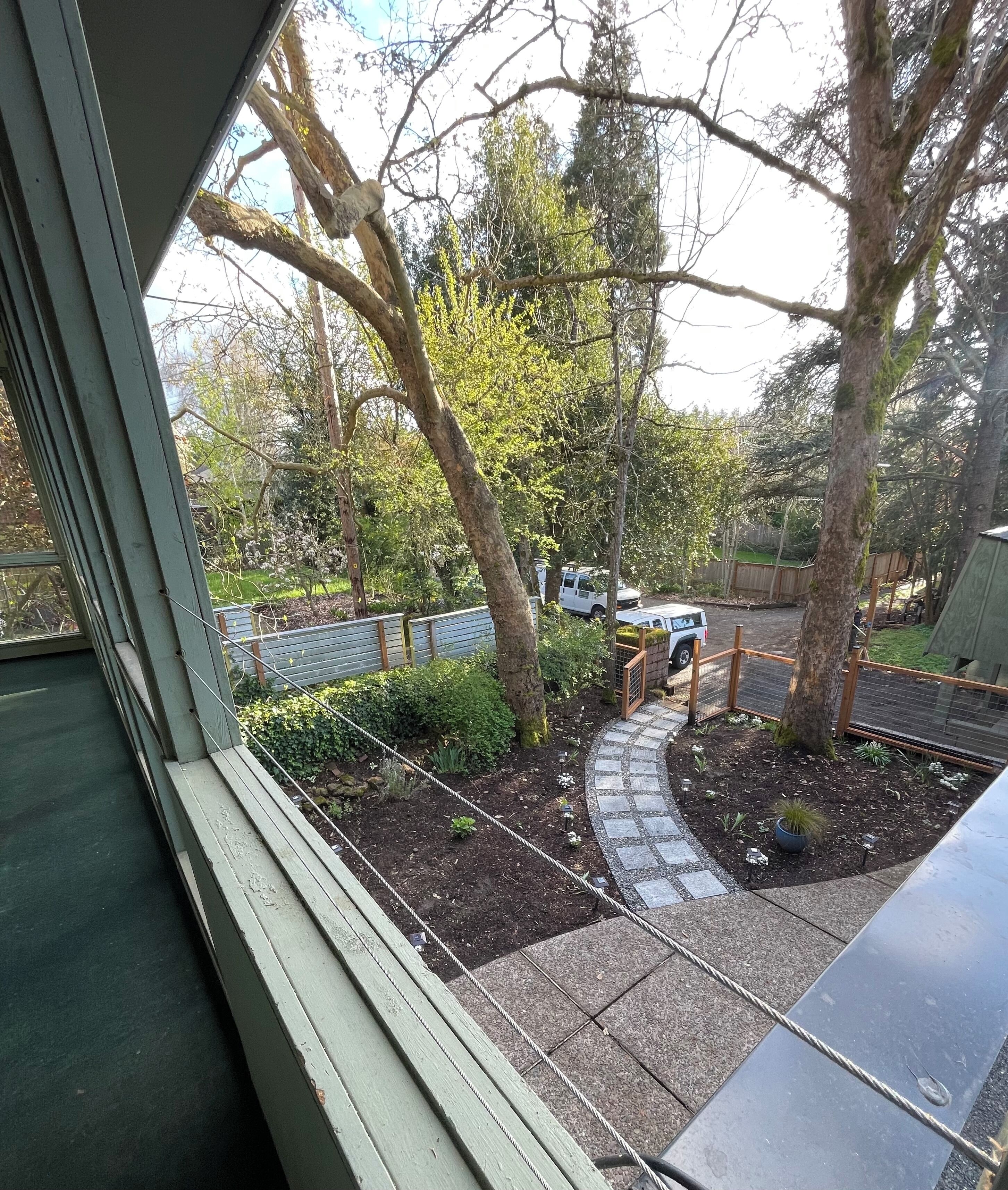 Une vue depuis une fenêtre donnant sur un jardin avec un chemin pavé, des arbres et un véhicule garé.