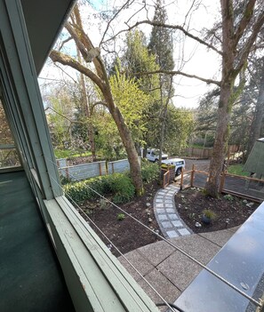 Vista desde una ventana a un jardín con un camino empedrado, árboles y un vehículo estacionado.