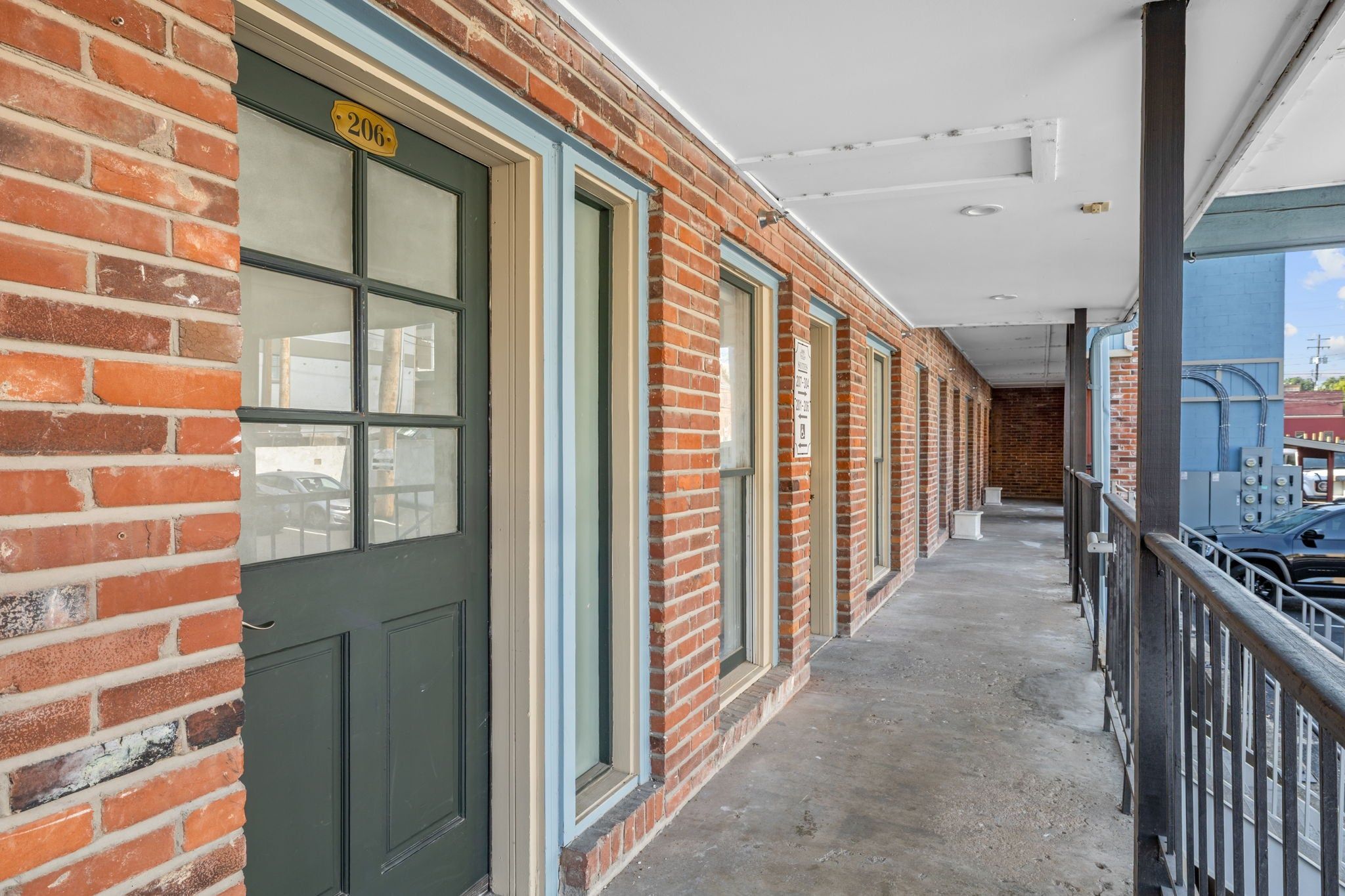 Un couloir à l’extérieur en briques, une porte portant l’inscription « 206 », et un balcon avec une rambarde en métal.