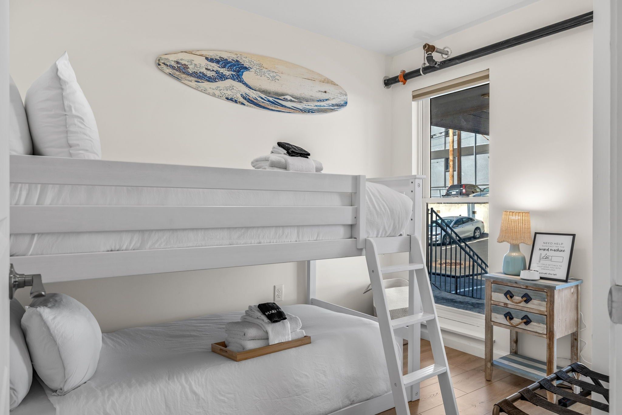 Un lit superposé avec une échelle, une table de chevet avec une lampe et une vue sur un parking à travers une porte vitrée.