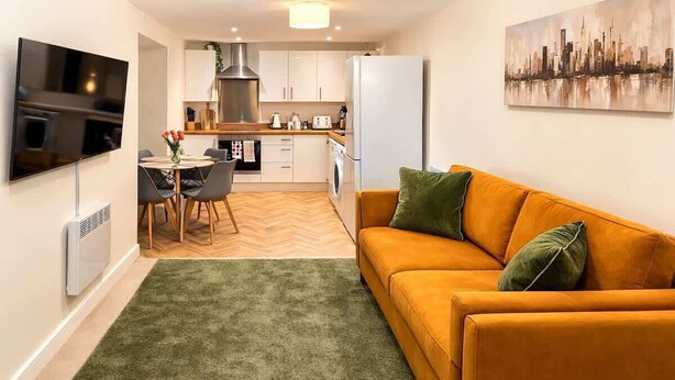 A compact living space with a sofa, a dining table with chairs, a kitchen area, and a flat-screen TV mounted on the wall. - Stylish Central Penthouse: Free Parking & Wi-Fi (Derby)
