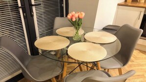 A dining area with a glass table surrounded by grey chairs, a vase with flowers, and a window with blinds. - Stylish Central Penthouse: Free Parking & Wi-Fi (Derby)