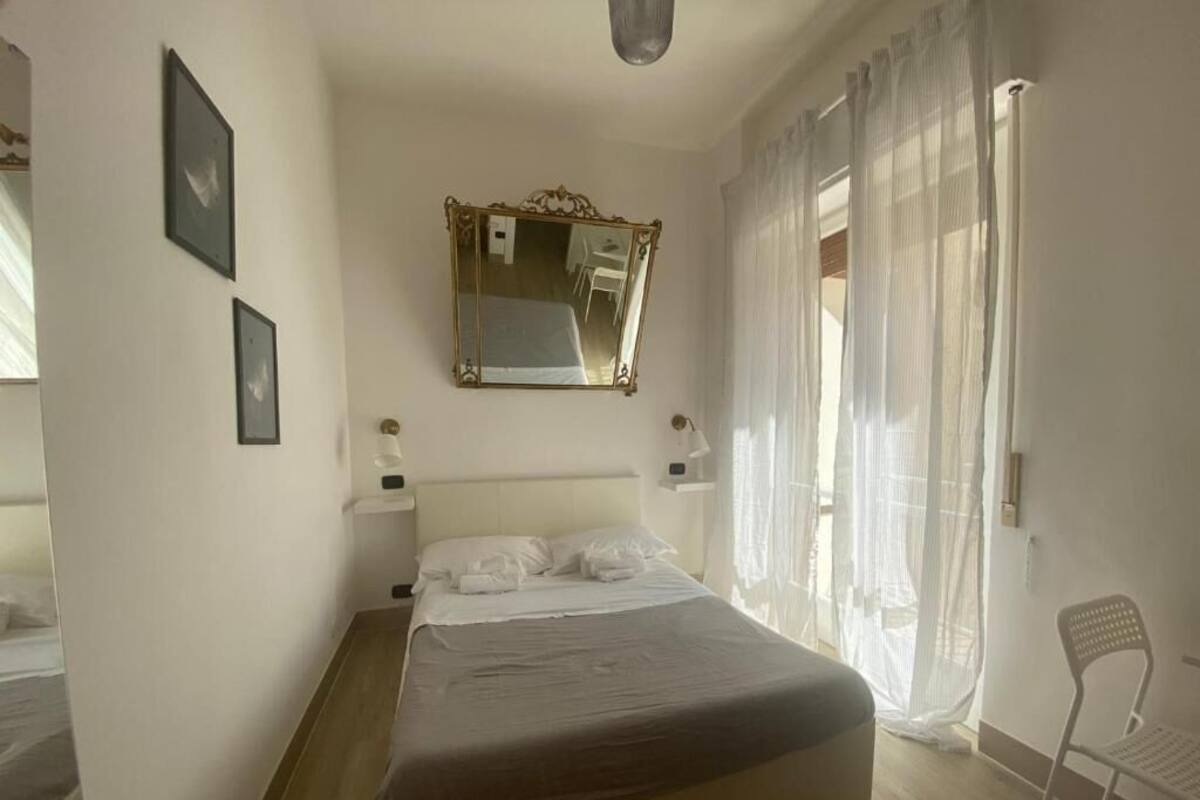 Un dormitorio con cama, espejo, silla y ventana con cortinas traslúcidas.