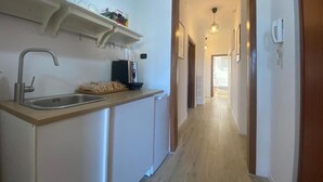A narrow hallway with a kitchen area on the left, featuring a sink, a coffee machine, and a shelf with various items.