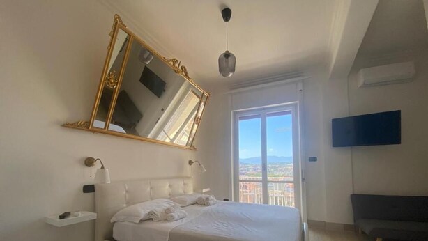 A bedroom with a bed, a large mirror, a bedside table, a TV, and a balcony with a view.