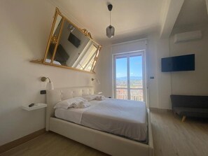 A bedroom with a bed, a large mirror, a bedside table, a TV, and a balcony with a view. - b&b 10 mt dal duomo (Benevento)