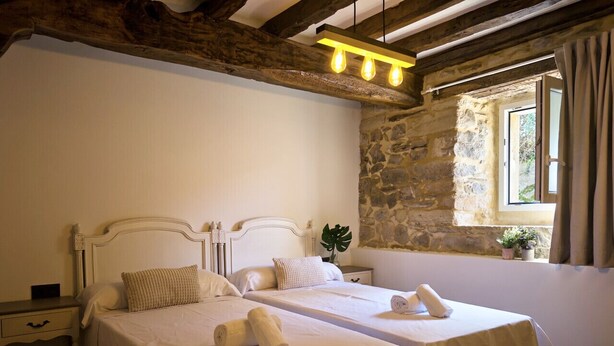 A double room with two beds, a stone wall, a window with curtains, and a wooden ceiling. - Casa De Postas - Bajo con jardín, 2 dormitorios (San Mamés de Meruelo)