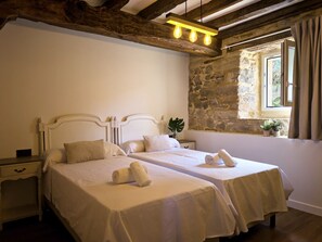 A double room with two beds, a stone wall, a window with curtains, and a wooden ceiling. - Casa De Postas - Bajo con jardín, 2 dormitorios (San Mamés de Meruelo)