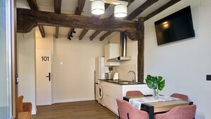 A modern kitchen with wooden beams, a dining area with pink chairs, and a TV mounted on the wall. - Casa De Postas - Bajo con jardín, 2 dormitorios (San Mamés de Meruelo)