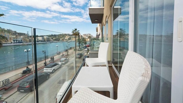 Ein Balkon mit weißem Korbmöbel, der auf einen Wasserblick mit geparkten Autos und einem Fußweg blickt.