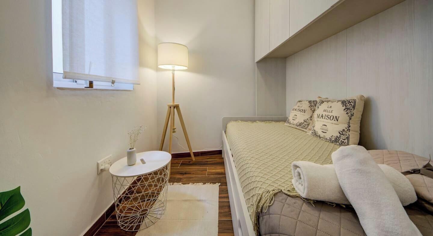 A bedroom with a bed, a standing lamp, a small round table with a vase, and a window with a blind.