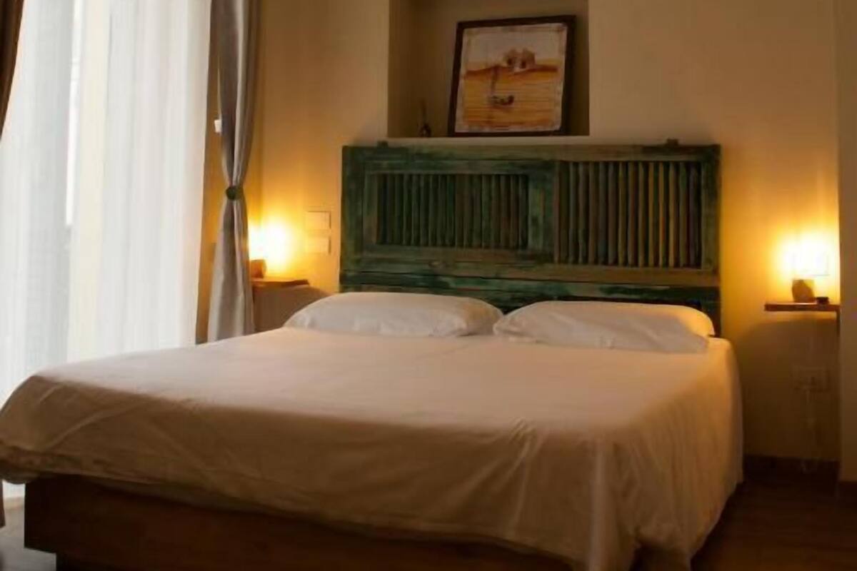 Un dormitorio con una cama, cabecera, un cuadro enmarco y dos lámparas de noche.